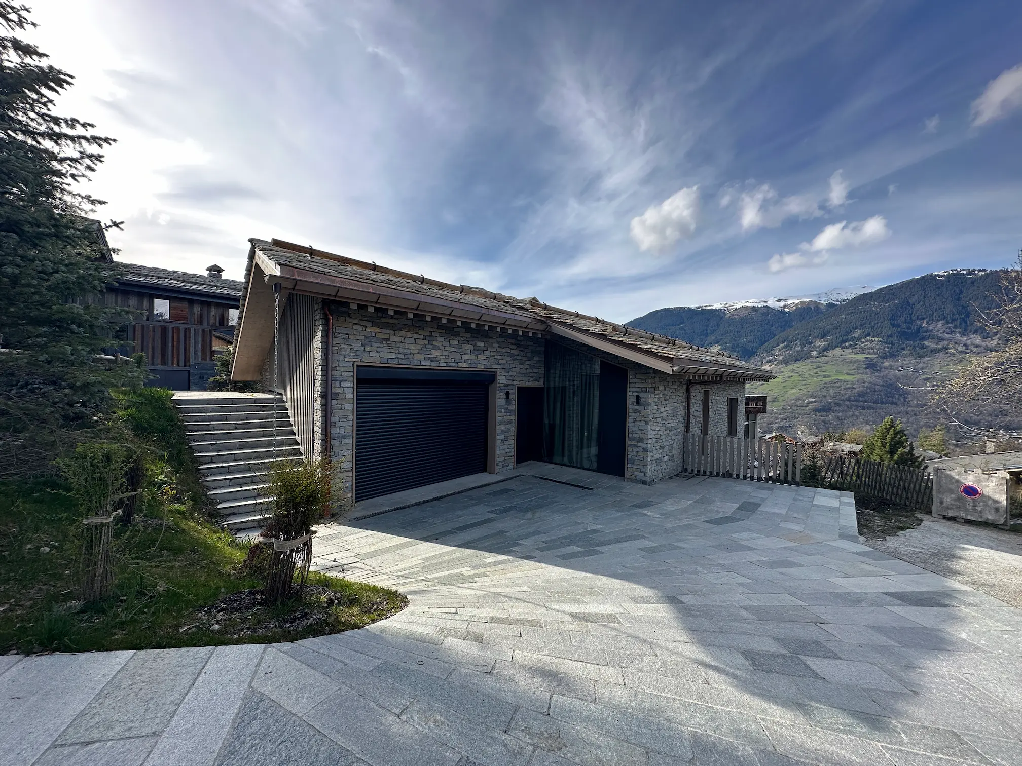 Maison en pierre avec garage, terrasse pavée et vue sur les montagnes enneigées sous un ciel dégagé